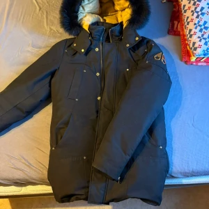 Svart dunjacka från Moose Knuckles - Säljer en svart dunjacka från Moose Knuckles med guldfärgade detaljer och gul vadderad insida. Jackan har avtagbar huva med svart päls, flera fickor och dragkedja framtill. Perfekt för kalla dagar och riktigt snygg streetstil.