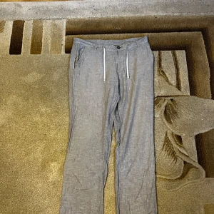 Grå linnebyxor från Livergy med snörning - Snygga grå linnebyxor från Livergy med raka ben och vita snören i midjan. Byxorna har sidfickor, bälteshällor och knäppning fram. Perfekta för en chill och avslappnad stil under varmare dagar.