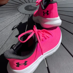 Rosa sneakers från Under Armour - Snygga och sportiga sneakers från Under Armour i stark rosa färg med svart logga och detaljer. Skorna har snörning, vit platt sula och ventilerande meshmaterial. Perfekta för dig som gillar att sticka ut och vill ha bekväma skor till träning eller vardag. Aldirg använda 10/10 skick nypris 699kr