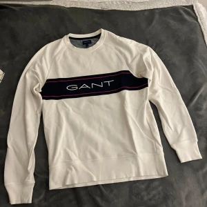 Vit sweatshirt från GANT med logga - Vit sweatshirt från GANT med klassisk rund hals och lång ärm. Tröjan har en bred marinblå rand över bröstet med GANT-loggan i vitt och röda detaljer. Tillverkad i mjuk bomull, perfekt för en clean och stilren look.
