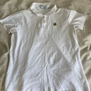 Lyle & Scott vit piké herr - Vit pikétröja från Lyle & Scott med gul logga på bröstet. Tröjan har krage och två knappar framtill.