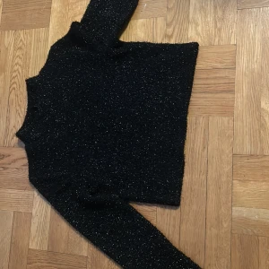 Svart glittrig polotröja Divided - Svart polotröja från Divided med glittrigt material som ger en snygg shimmer-effekt. Tröjan har lång ärm och en tight passform, perfekt för att sticka ut lite extra. Materialet är mjukt och stretchigt, vilket gör den bekväm att bära.