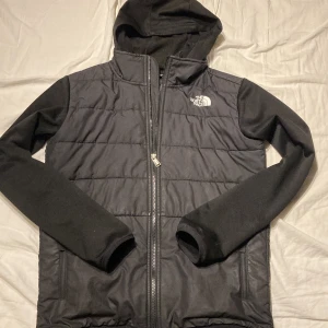 Svart cardigan/hybridjacka från The North Face - Svart hybridjacka från The North Face med huva och långa ärmar i mjukt material. Jackan har quiltad framsida, dragkedja hela vägen och två sidofickor med dragkedja. Perfekt för kyliga dagar och snygg streetstil. Passar för ungefär xs-s. Skriv om ni har frågor och skicka gärna bud!
