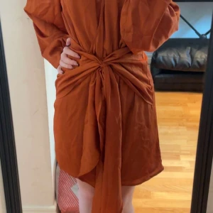 Rostbrun Gina Tricot klänning  - Superfin rostbrun kortklänning från Gina Tricot med långa ärmar, v-ringning och snygg omlottknytning i midjan. Klänningen har en loose fit och är gjord i ett mjukt, lätt material som känns skönt mot huden. Perfekt för dig som gillar trendiga och bekväma plagg.
