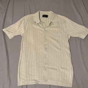 Beige ribbad skjorta från ASOS Design - Snygg beige skjorta från ASOS Design i ribbad bomull. Skjortan har korta ärmar, klassisk krage och knäppning framtill. Perfekt för dig som gillar en clean och stilren look med lite retrovibbar.