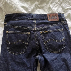 Mörkblå bootcut jeans från Lee - Snygga mörkblå bootcut jeans från Lee, modell Felton Flare. Klassisk femficksdesign med kontrastsömmar och Lee-lapp bak i midjan. Tillverkade i 100% bomull för en autentisk jeanskänsla. Perfekta för dig som gillar retrovibbar och bredare ben. Passar mig bra i längden som är 172 men små i midjan, skulle säga att de motsvarar 26/27
