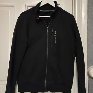 Svart ziptröja från Sail Racing - Svart tröja från Sail Racing med hel dragkedja och en bröstficka med logga. Tröjan har hög krage, ribbade muddar och lång ärm. Perfekt för dig som gillar stilren och sportig look.