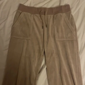 Juicy Couture byxor - Supermjuka beige mjukisbyxor från Juicy Couture i velour med bred resår och snörning i midjan. Stora fickor fram och bak med tryckknappar och broderad JC-logga på ena fickan. Perfekta för chill dagar och snyggt avslappnad look.