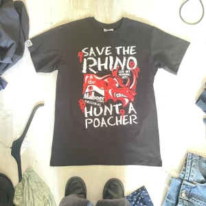 Svart t-shirt med tryck från Malboer - Svart t-shirt från Malboer i bomull med stort grafiskt tryck framtill i vitt och rött med texten 'Save the Rhino, Hunt a Poacher' och en röd noshörning. Klassisk passform och rund hals. Perfekt för dig som gillar statement-plagg.
