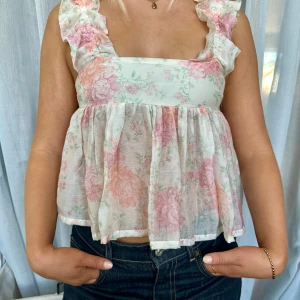 Blommig volangblus med knytband i nyskick - Supersöt blommig blus i ljusa pastellfärger med volangprydda axelband och knytband i ryggen i nyskick. Luftig och flowy modell med fyrkantig ringning och romantiskt mönster i rosa, grön och vit. Perfekt för sommarens alla tillfällen!