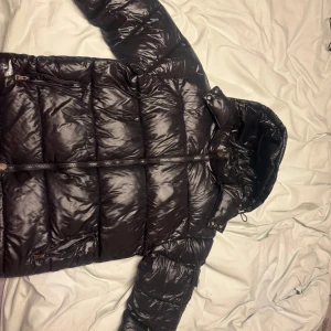 Moncler maya  - Säljer en svart pufferjacka från Moncler med glansig finish och huva. Jackan har dragkedja framtill, flera fickor med dragkedja och Moncler-logga på ärmen. Perfekt för kalla dagar när du vill hålla stilen och värmen. ett litet hål vid höger fickan har tejpat över de så de syns ej men går nog lösa bättre