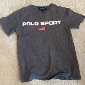 Grå Polo Sport t-shirt från Ralph Lauren - Mörkgrå t-shirt från Polo Sport Ralph Lauren med vit och röd logga framtill samt amerikansk flagga. Klassisk rund hals och korta ärmar. Skön och mjuk bomullskvalitet, perfekt för en avslappnad stil.