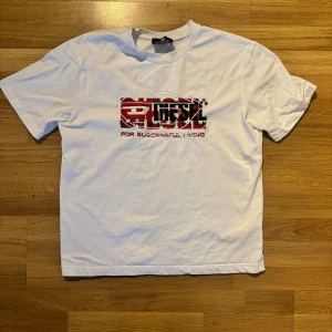 Vit t-shirt med röd Diesel-logga - Vit t-shirt från Diesel med broderad röd och svart logga framtill och texten 'For Successful Living'. Klassisk passform, rund hals och korta ärmar. Tillverkad i 100% polyester för en lätt och skön känsla.