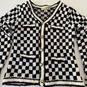 Rutig kofta i svart och vitt från e-woman - Säljer en fluffig kofta från e-woman med svartvitt rutigt mönster, guldiga detaljer och knappar framtill. Koftan har fyra fickor och v-ringad hals. Perfekt för dig som vill sticka ut med en cool och trendig look.