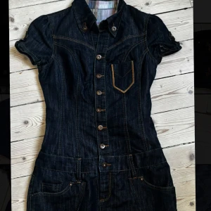 Figursydd jeansklänning med broderi - Snygg mörkblå jeansklänning med kort ärm, knappar hela vägen framtill och coola fickor både fram och bak. Klänningen har orange broderi på bakfickorna och markerade sömmar som ger en schysst denimlook. Perfekt för dig som gillar street och denim vibes.