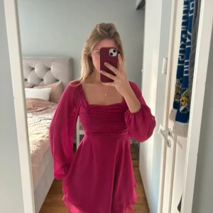 Rosa kortklänning med puffärmar - Supersöt rosa kortklänning med långa puffärmar och rynkade detaljer över bysten. Klänningen har en flowy kjol och fyrkantig ringning. Perfekt för dig som vill sticka ut med en färgstark look!