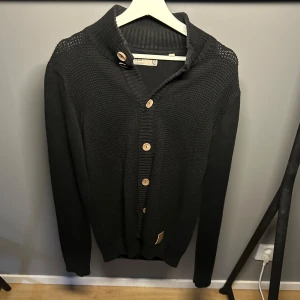 Svart stickad kofta med träknappar - Svart stickad kofta med grovstickad struktur och ribbad krage. Stängs med stora träfärgade knappar framtill. Långärmad och perfekt för lager-på-lager. Enkel och stilren design som funkar till det mesta.