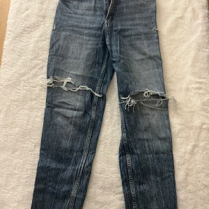 Blå slitna jeans med hål vid knäna - Säljer ett par blå jeans med coola slitningar och stora hål vid båda knäna. Jeansen har klassisk femficksdesign och rak passform. Perfekta för dig som gillar en avslappnad och trendig streetstil.