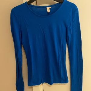 Blå långärmad topp från Nelly - En enkel och stilren blå långärmad topp från Nelly. Modellen är figurnära med rund halsringning och tillverkad i ett mjukt, stretchigt material som känns skönt mot huden. Perfekt att styla med jeans eller kjol för en clean look.