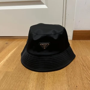 Svart bucket hat från Prada - Stilren svart bucket hat från Prada med klassisk siluett och ikonisk metallogo framtill. Hatten är tillverkad i glansig syntet och har bred, nedåtlutande brätte. Perfekt accessoar för att lyfta din outfit med en lyxig touch.