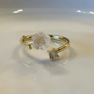 Guldfärgad ring med ros och sten - Unik guldfärgad ring med öppen design, dekorerad med en genomskinlig ros i akryl och en gnistrande klar sten. Ringen har små blad-detaljer på sidorna som ger en romantisk och trendig känsla. Perfekt accessoar för dig som gillar detaljer och blommiga inslag.