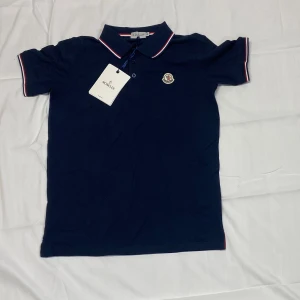 Marinblå Moncler pikétröja M - Snygg marinblå pikétröja från Moncler i bomull. Klassisk krage med rödvita detaljer på krage och ärmslut samt Moncler-logga på bröstet. Perfekt för dig som gillar stilren och sportig look.