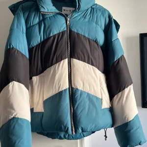 Färgglad pufferjacka från NA-KD - Säljer en snygg pufferjacka från NA-KD med breda blockränder i turkos, svart och offwhite. Jackan har dragkedja framtill, huva och dragsko nertill för extra stilpoäng. Perfekt för kalla dagar när du vill sticka ut lite extra. Jackan är även i storlek 36( s-m)