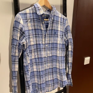 Blårutig skjorta från Ralph Lauren - Snygg blårutig skjorta från Ralph Lauren i bomull med klassisk krage och knappar framtill. Custom fit-modell med broderad logga på bröstet. Perfekt för dig som gillar en stilren och avslappnad look. Storlek 14-16 år