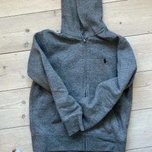 Grå hoodie från Polo Ralph Lauren - Snygg grå hoodie från Polo Ralph Lauren med dragkedja och klassisk huva. Tröjan har en liten mörkblå logga broderad på bröstet och fickor framtill.