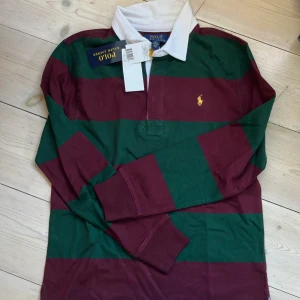 Randig pikétröja Polo Ralph Lauren - Säljer en klassisk långärmad pikétröja från Polo Ralph Lauren med breda vinröda och gröna ränder, vit krage och gul broderad logga på bröstet. Tröjan är i bomull. 