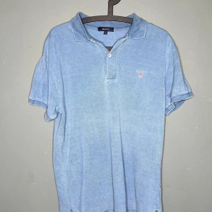 Blå pikétröja från GANT i bomull - Klassisk ljusblå pikétröja från GANT i storlek L. Tröjan har korta ärmar, krage och knäppning framtill samt broderad GANT-logga på bröstet. Tillverkad i mjuk bomull för en skön och avslappnad stil.