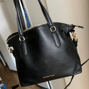 Svart axelväska från Valentino - Snygg svart axelväska från Valentino i strukturerat skinn med guldfärgade detaljer och dragkedja. Väskan har både handtag och bred axelrem med Valentino-logga i nitar. Rymligt huvudfack och innerficka för småsaker. Perfekt för dig som vill ha en stilren och trendig väska.