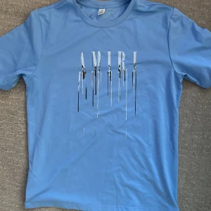 Ljusblå Amiri t-shirt med tryck - Snygg ljusblå t-shirt från Amiri med korta ärmar och coolt tryck framtill där loggan ser ut att smälta ner. Skön passform och tillverkad i mjuk bomull. Perfekt för dig som vill sticka ut med en unik streetstyle.