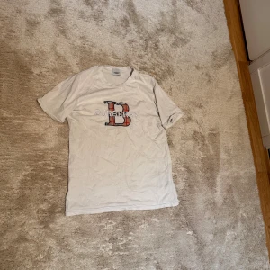 Vit Burberry t-shirt med logga - Vit kortärmad t-shirt från Burberry med stor B-logga och text på bröstet. Tillverkad i en mjuk blandning av bomull och polyester med lite lycra för stretch. Klassisk passform och rund halsringning, perfekt för en clean och stilren look.