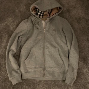 Grå hoodie från Burberry med dragkedja - Säljer en grå hoodie från Burberry med klassiskt rutigt foder i huvan. Tröjan har dragkedja framtill, snörning i huvan och två stora fickor. Perfekt för dig som vill ha en stilren och lyxig look med ikoniska Burberry-detaljer.