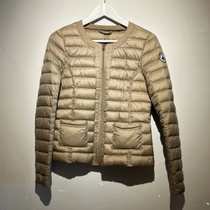 Beige dunjacka från JOTT - Snygg beige dunjacka från JOTT med quiltad design och rund halsringning. Jackan har två stora fickor framtill, dragkedja och ett broderat JOTT-märke på ärmen. Lätt och smidig modell som passar perfekt till kyliga dagar. Köpt på Jackie butiken.