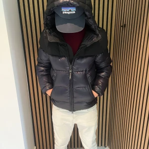 Svart och mörkblå pufferjacka Replay - Säljer en fet pufferjacka från Replay i svart och mörkblått. Jackan har huva med tryck, dragkedja framtill och cool device pocket på insidan. Perfekt för kalla dagar när du vill hålla stilen och vara varm. Oversized känsla och riktigt skön passform.