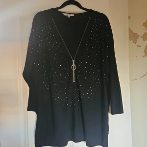 Svart tröja med dragkedja från Cellbes - Svart långärmad tröja från Cellbes med snygga silvriga nitar framtill och en dekorativ dragkedja med ring och tofs. Tröjan har en avslappnad passform och passar perfekt när du vill ha något stilrent men ändå med lite edge.