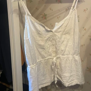 Vit linnetopp med knyt från frk. - Säljer en vit linnetopp från frk. med smala justerbara axelband, knappar framtill och knyt i midjan. Toppen har v-ringning och lätt rynkade detaljer samt en luftig passform, perfekt för varma dagar.
