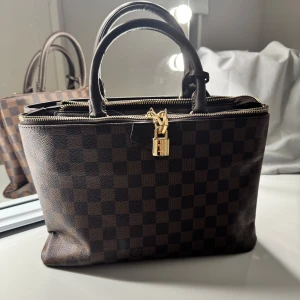 Handväska brun - Säljer en klassisk handväska i brunt Damier-mönster med guldiga metalldetaljer och lås. Väskan har både handtag och avtagbar axelrem, dragkedja upptill och är tillverkad i skinn. Perfekt för dig som gillar lyxiga accessoarer med ikonisk design. Den är köpt utomlands alltså inget speciellt märke. 