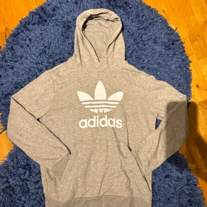 Grå Adidas hoodie - Säljer en grå hoodie från Adidas med stor vit adidas logga på bröstet. Klassisk modell med huva och känguruficka framtill. Mjuk och skön i bomullsblandning, perfekt för chill eller träning. Långärmad och med ribbade muddar för extra komfort.