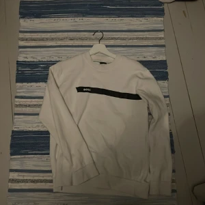 Vit sweatshirt från BOSS med svart detalj - Vit sweatshirt från BOSS med svart band och logga över bröstet. Klassisk rund halsringning och långa ärmar. Perfekt för dig som gillar stilren och enkel design med en touch av streetkänsla.