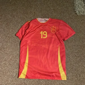 Röd Spanien fotbollströja Lamine Yamal - Säljer en röd Spanien fotbollströja med gula detaljer och nummer 19, Lamine Yamal på ryggen. Tröjan har korta ärmar, rund hals och är tillverkad i ett lätt, syntetiskt material som andas. Perfekt för dig som vill sticka ut på planen eller på läktaren.