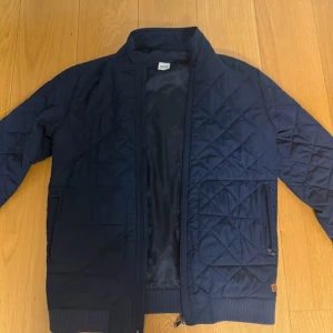 Marinblå quiltad jacka från Hugo Boss - Snygg marinblå quiltad jacka från Hugo Boss med dragkedja framtill, ribbade muddar och två fickor med dragkedja. Jackan har ett diskret mönster och innerfoder med Boss-logga. Perfekt för dig som vill ha en stilren och modern look.