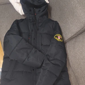 Svart pufferjacka Zavetti Canada XL jackor  - Svart pufferjacka från Zavetti Canada i storlek XL. Jackan har huva med snörning, flera fickor och dragkedja framtill. Snyggt broderat märke på ärmen och tryckt logga på fodret. Perfekt för kalla dagar med sin rejäla och vadderade känsla.