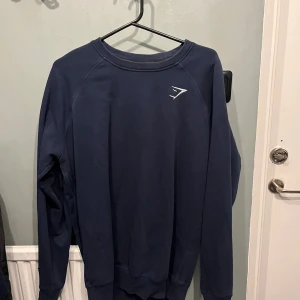 Mörkblå sweatshirt från Gymshark - Mörkblå sweatshirt från Gymshark med rund halsringning och diskret logga på bröstet. Tröjan har långa ärmar och ribbade muddar vid hals, ärmslut och nederkant. Perfekt för chill eller träning. Finns en liten skada på armen 
