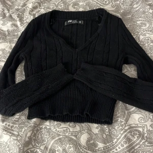 Svart ribbad stickad tröja New Yorker  - Svart stickad tröja från New Yorker i storlek XS. Tröjan är croppad med v-ringning, ribbade muddar och lång ärm. Snyggt kabelstickat mönster som ger en klassisk vibe. 