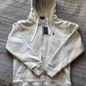 Grå hoodie från Polo Ralph Lauren - Snygg ljusgrå hoodie från Polo Ralph Lauren med dragkedja, huva och vita snören. Klassisk broderad logga i marinblått på bröstet. Mjuk bomullsblandning och kängurufickor framtill. Perfekt för en chill och stilren look.