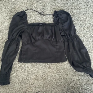 Svart blus med puffärmar från H&M Divided - Säljer en svart blus från H&M Divided med puffiga långa ärmar och smockdetaljer både fram och bak. Toppen har fyrkantig urringning och tunna knytband vid axlarna. Materialet är lätt och ger en snygg siluett, perfekt för att lyfta din outfit.