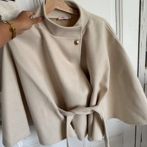 Beige kappa med bälte från Nelly - Slutsåld!! Stilren beige kappa från Nelly med hög krage, knytband i midjan och en snygg guldig knapp framtill. Aldrig använd och helt ny i super skick💗